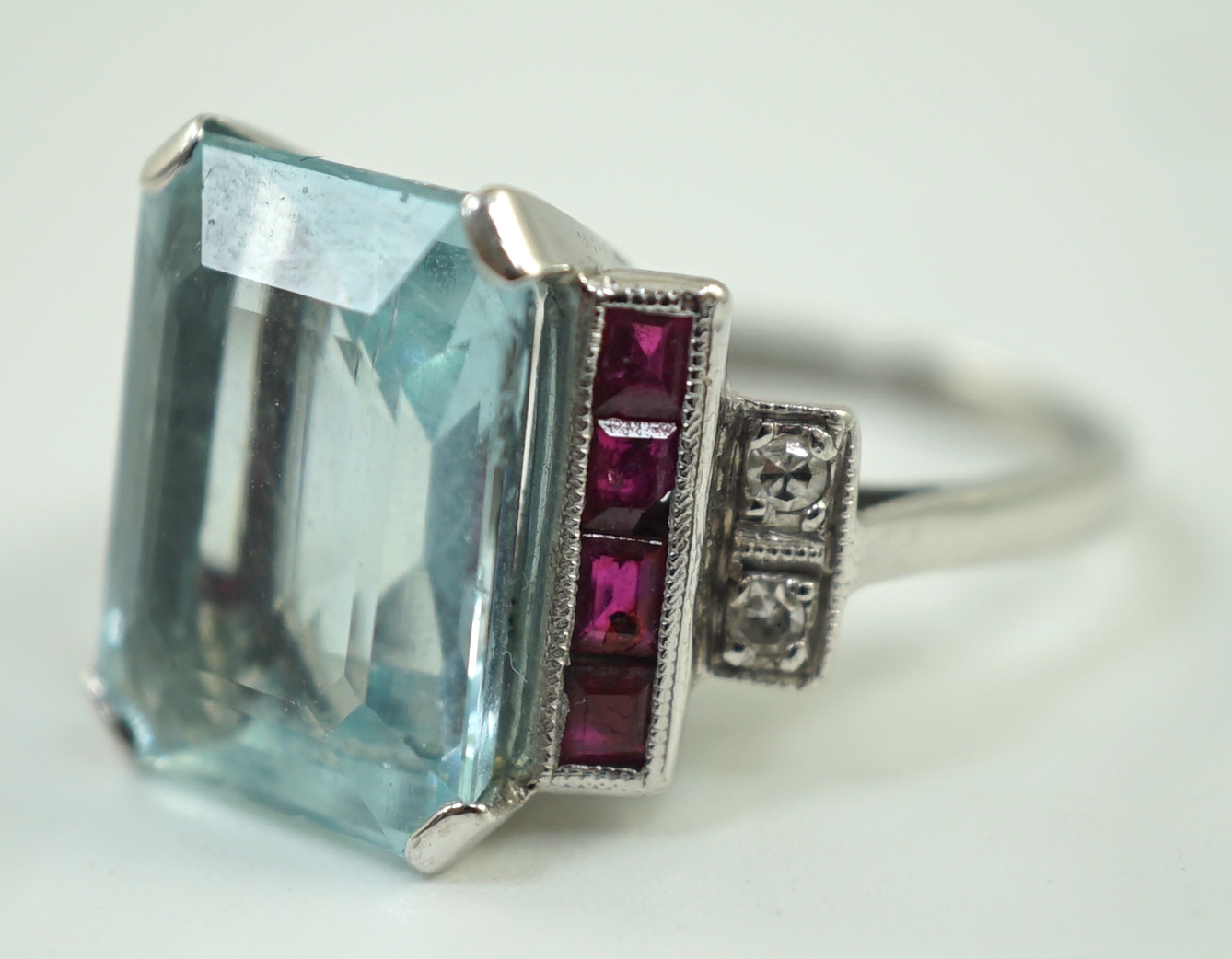 A platinum and single stone emerald cut aquamarine set dress ring, with ruby and diamond set stepped shoulders